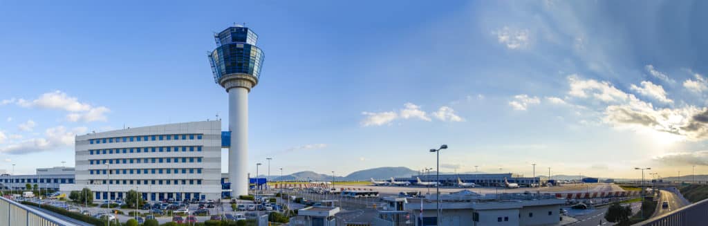 Panoramic view of an airport with a tall control tower, adjacent office building, runways, and mountains in the background under a blue sky with scattered clouds, highlighting the focus on airport customer experience.