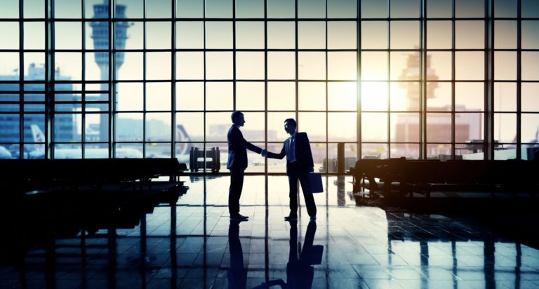 Two businesspeople shaking hands in an airport terminal with large windows, airplanes outside, and sunlight streaming in—capturing the spirit of Airport Leadership and Management Training through professional silhouettes and reflective floors.