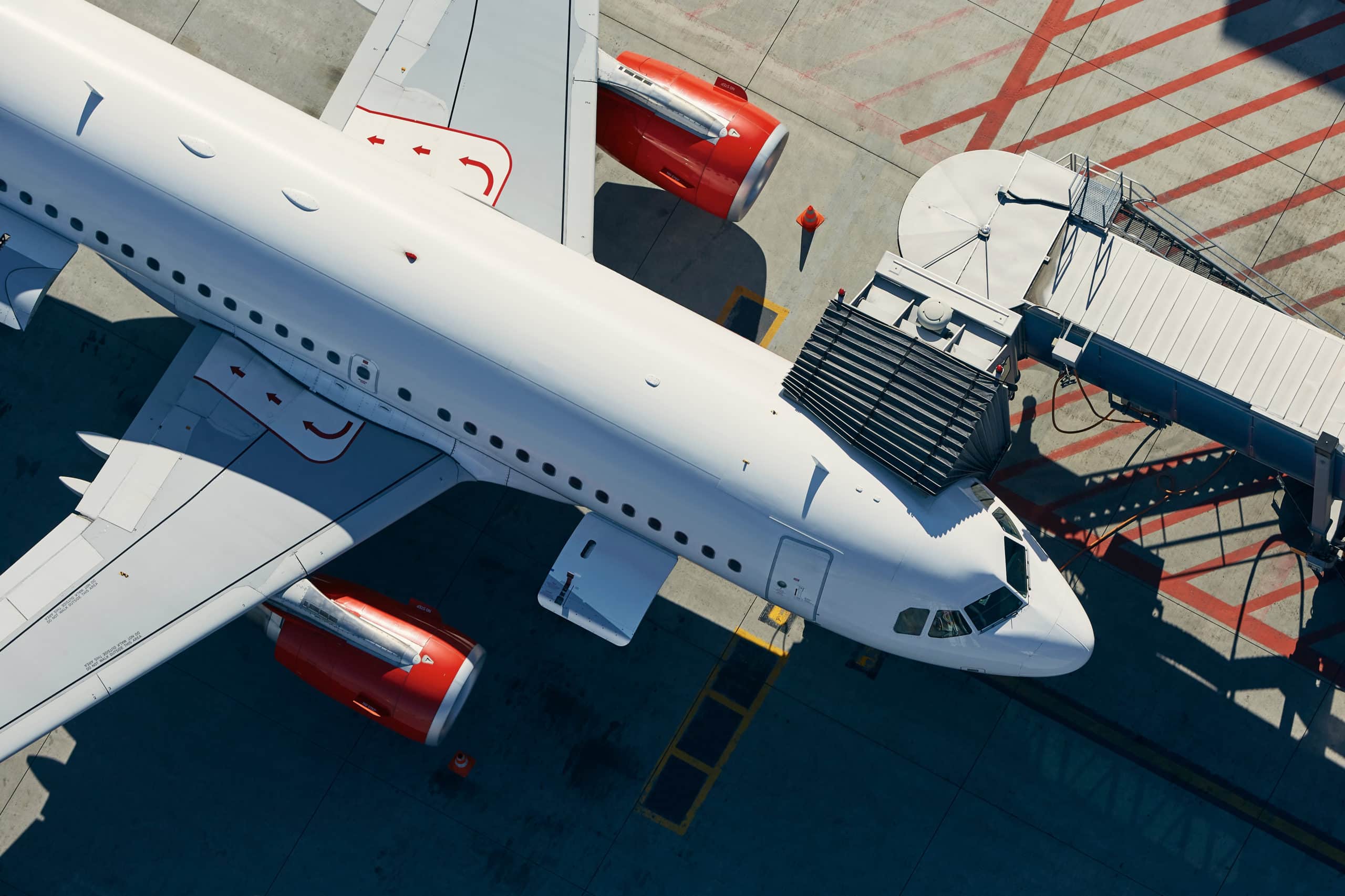 Aerial view of a white commercial airplane with red engines parked at an airport gate, connected to a jet bridge on the tarmac with marked lines.