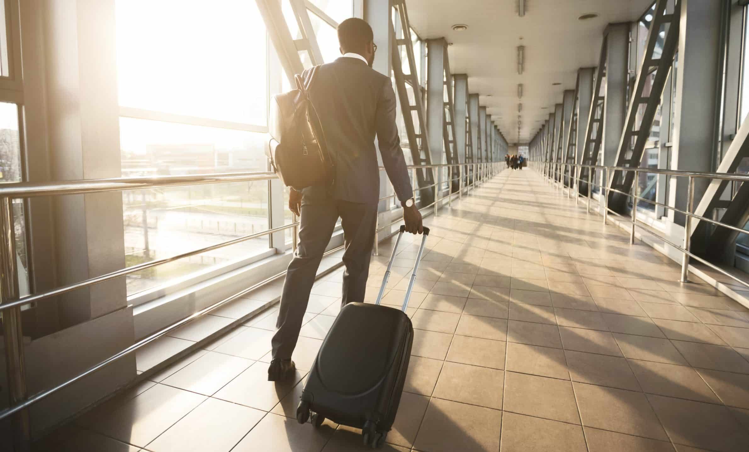 A man in a suit walks down a sunlit airport corridor, pulling a rolling suitcase with one hand and carrying a backpack, heading toward a distant group of people.