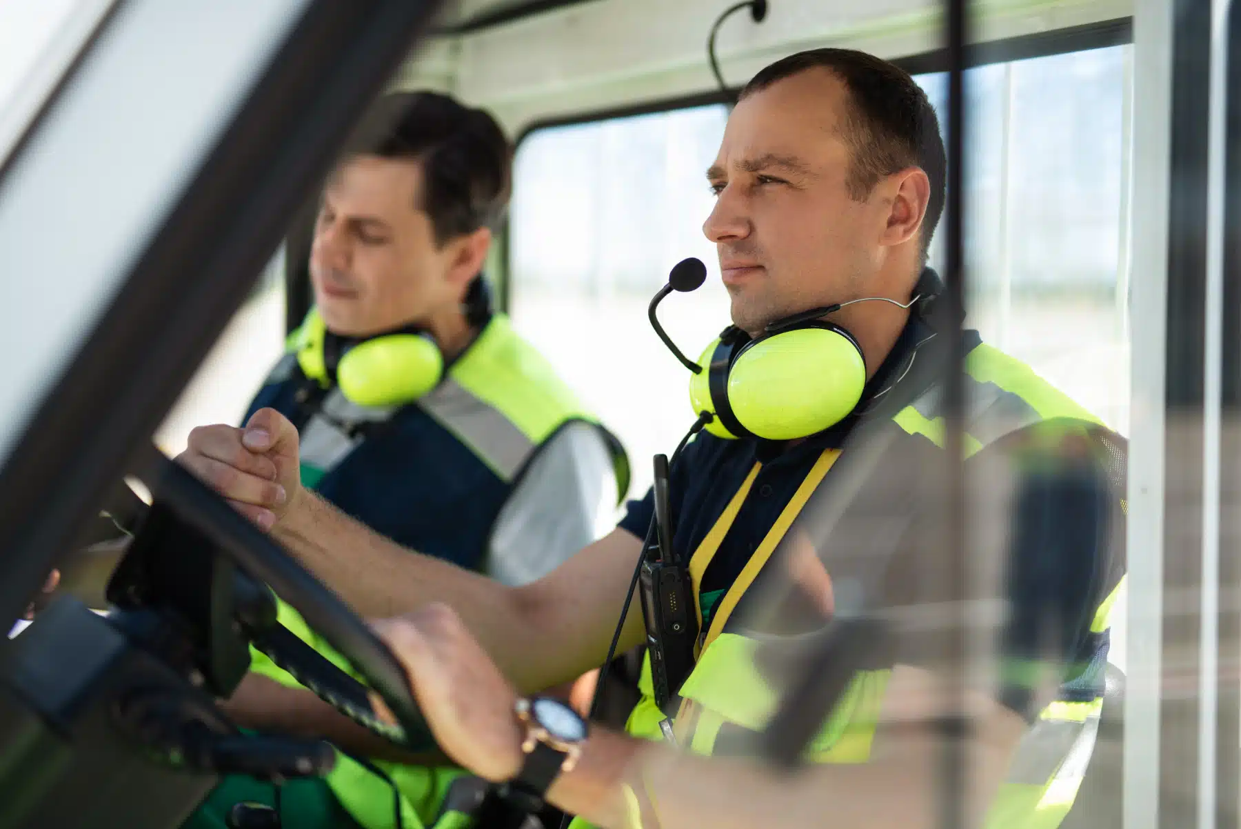 Two workers in high-visibility vests and headsets sit in a vehicle cab. The man in the foreground holds the steering wheel and looks ahead, while the other looks down, both focused on their tasks.
