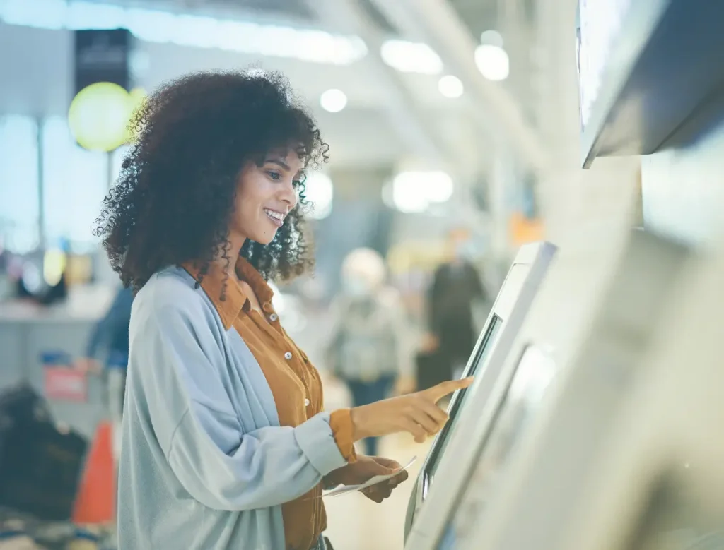 A woman with curly hair smiles while using a touchscreen kiosk at an airport, holding travel documents in one hand. The background shows a bright, busy terminal with people and signs.
