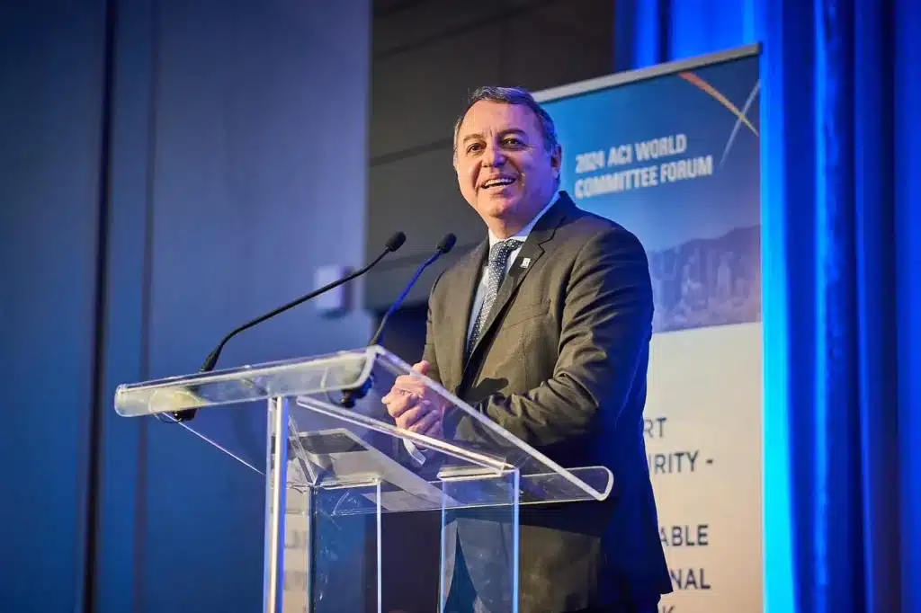A man in a suit speaks at a clear podium with two microphones, smiling. A blue and white banner behind him reads 2024 ACI WORLD COMMITTEE FORUM with additional partially visible text. Blue lighting illuminates the scene.
