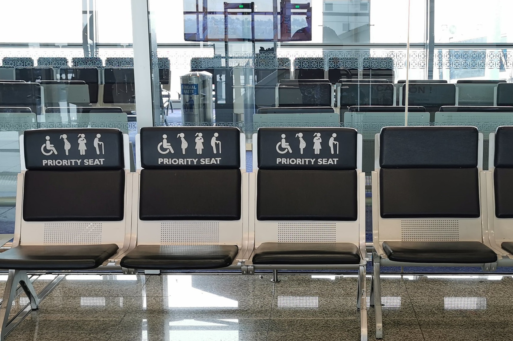 Four airport seats labeled Priority Seat with icons for people with disabilities, elderly, pregnant women, and adults with children highlight airport accessibility in a bright waiting area with glass walls.