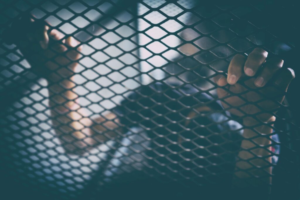 A person grips a metal wire fence with both hands, their face and body blurred in the background, evoking a somber scene of confinement—reminiscent of airport security areas where passenger facilitation is stalled. The lighting remains dim and moody.