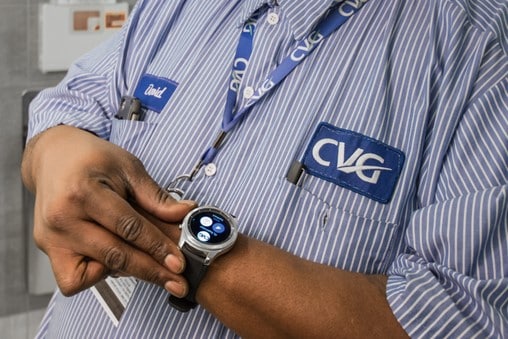 A person wearing a striped shirt with CVG logos and a name tag checks a smartwatch displaying various app icons, highlighting the role of Airport IT. The focus is on the hands and watch, not showing the person's face.