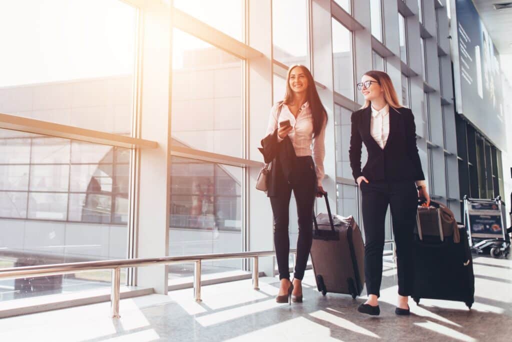 Two women in business attire walk through an airport terminal with rolling suitcases, smiling and talking. Sunlight streams through large windows, highlighting efficient passenger facilitation and luggage carts in the background.