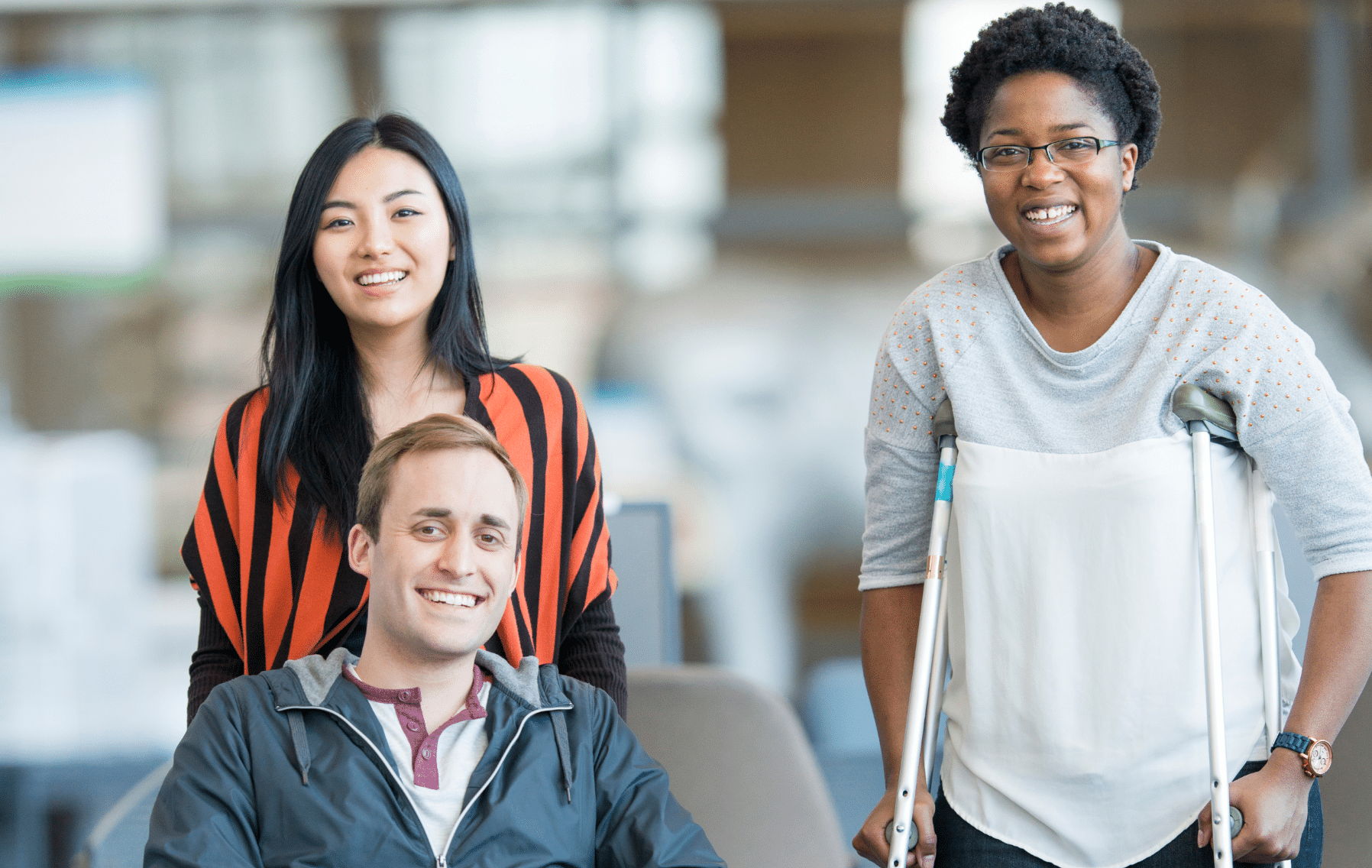 Three smiling people, one seated in a wheelchair, one with crutches, and one standing behind them, pose together in a bright indoor setting, highlighting the importance of Accessibility at airports for all travelers.