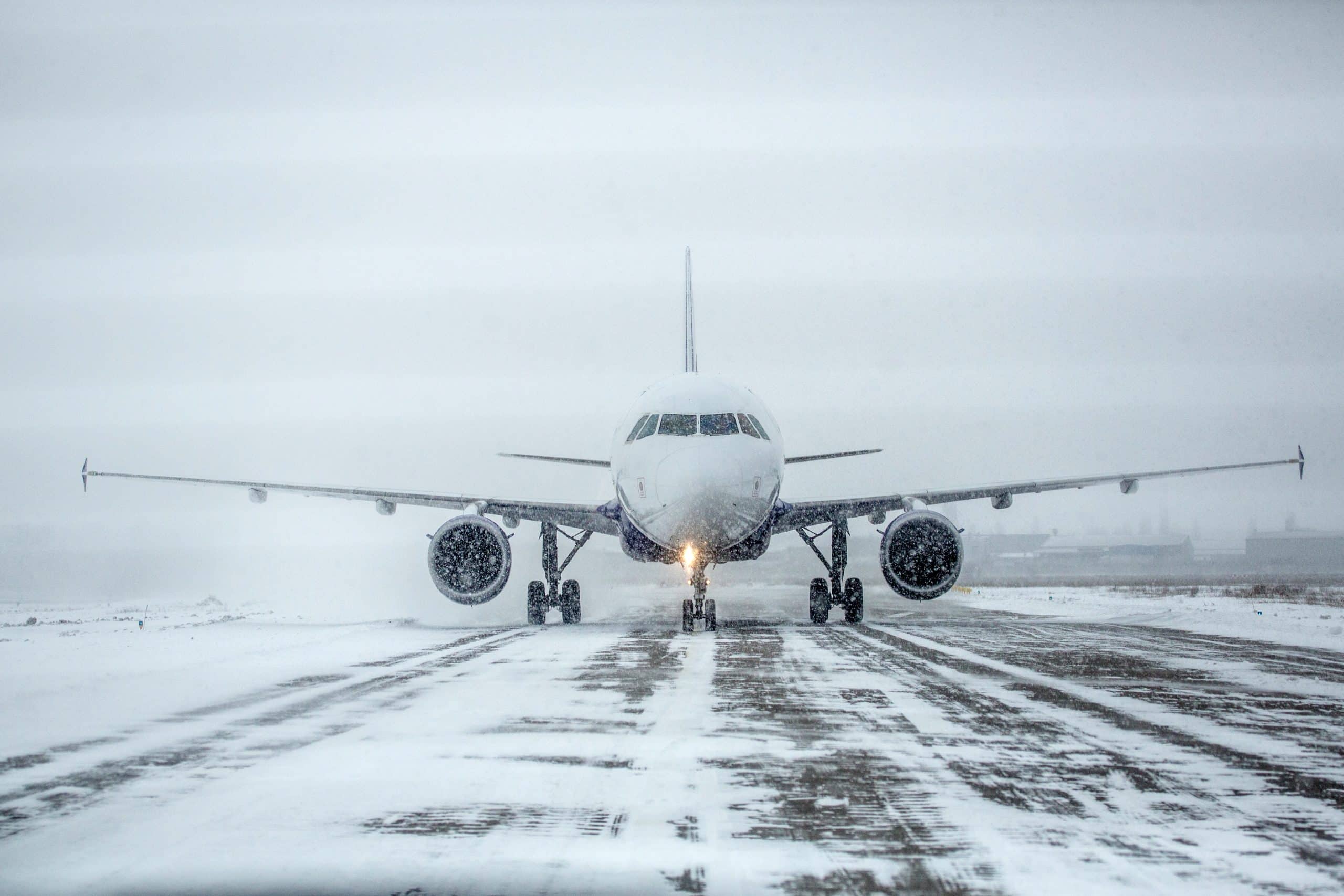 A commercial airplane is taxiing on a snow-covered runway at the airport during a snowstorm, with poor visibility and cloudy skies challenging passenger facilitation.