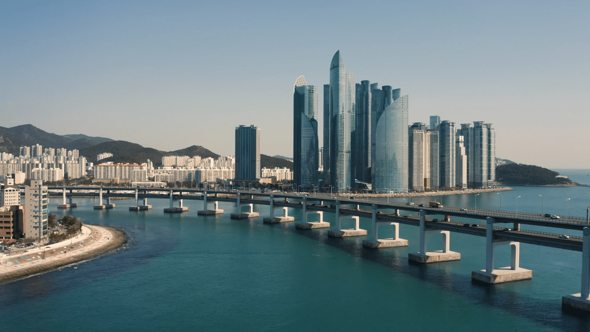 A cityscape featuring modern high-rise buildings along a coastline, with a bridge crossing over blue water in the foreground and mountains in the background under a clear sky.