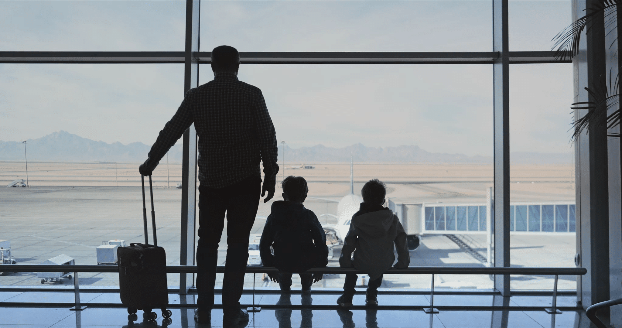 A man with a rolling suitcase stands beside two children sitting on a railing, all looking out a large airport window at a plane and runway with mountains in the distance—an image fit for the front page.