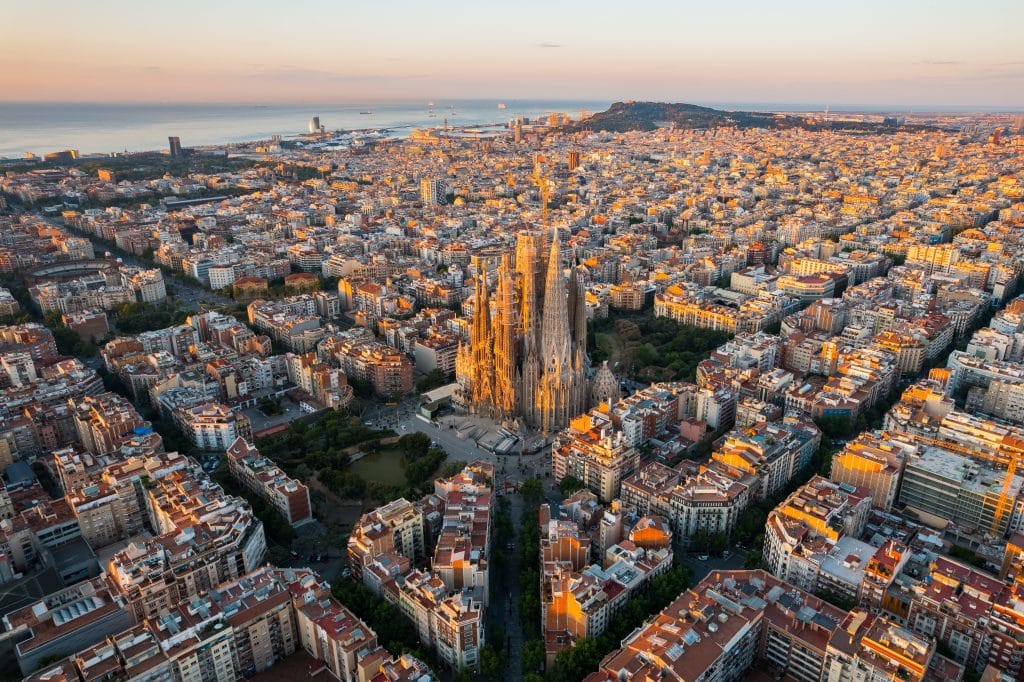 Aerial view of Barcelona at sunset, featuring the Sagrada Família basilica at the center, grid-like city blocks, and the Mediterranean Sea—capturing a city ready for change as Airports Innovate 2026 nears.