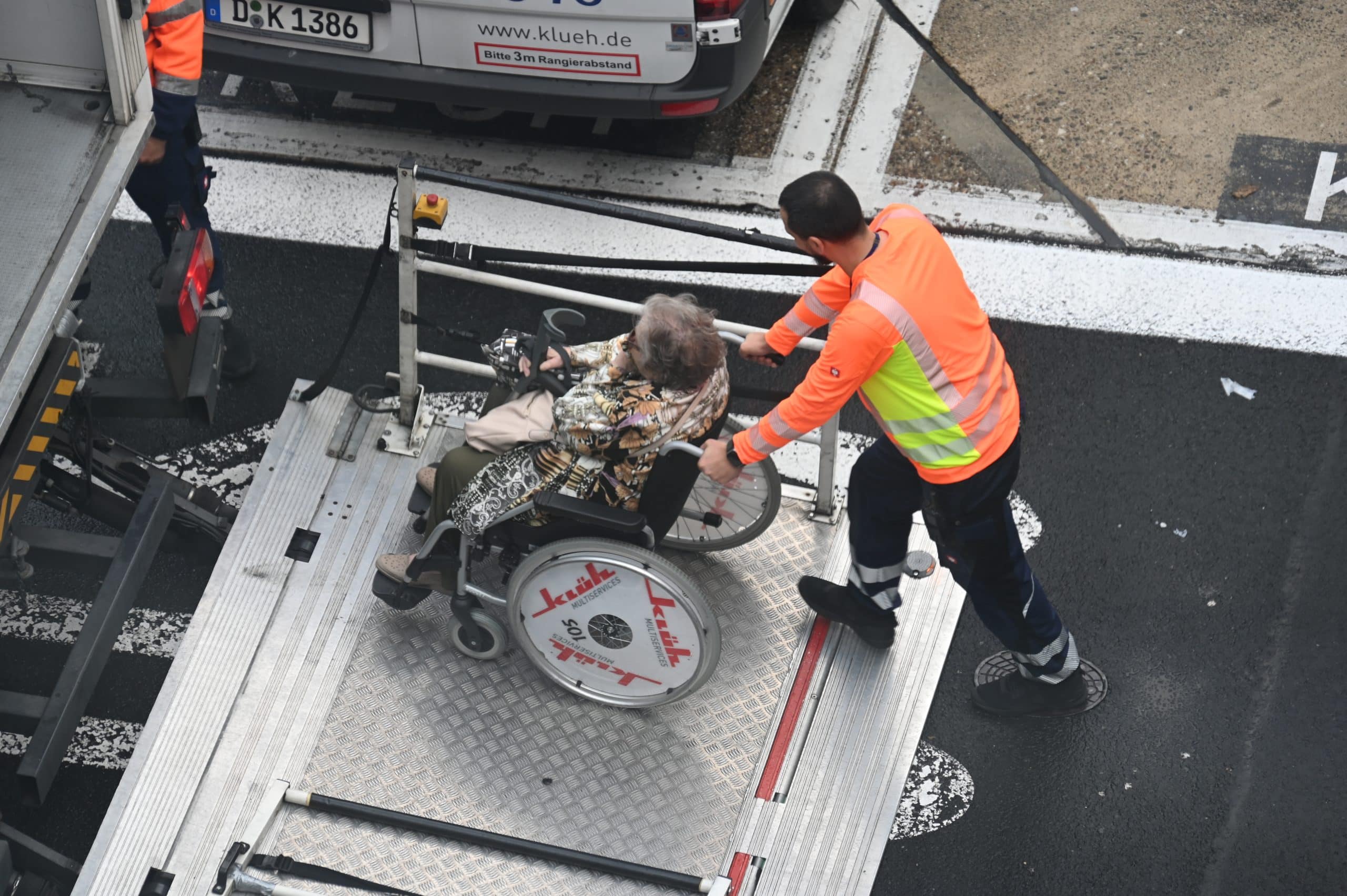 A person in a wheelchair is assisted onto an airport lift platform by a worker in a bright orange and yellow safety vest, highlighting airport accessibility as they prepare for boarding near an airplane.