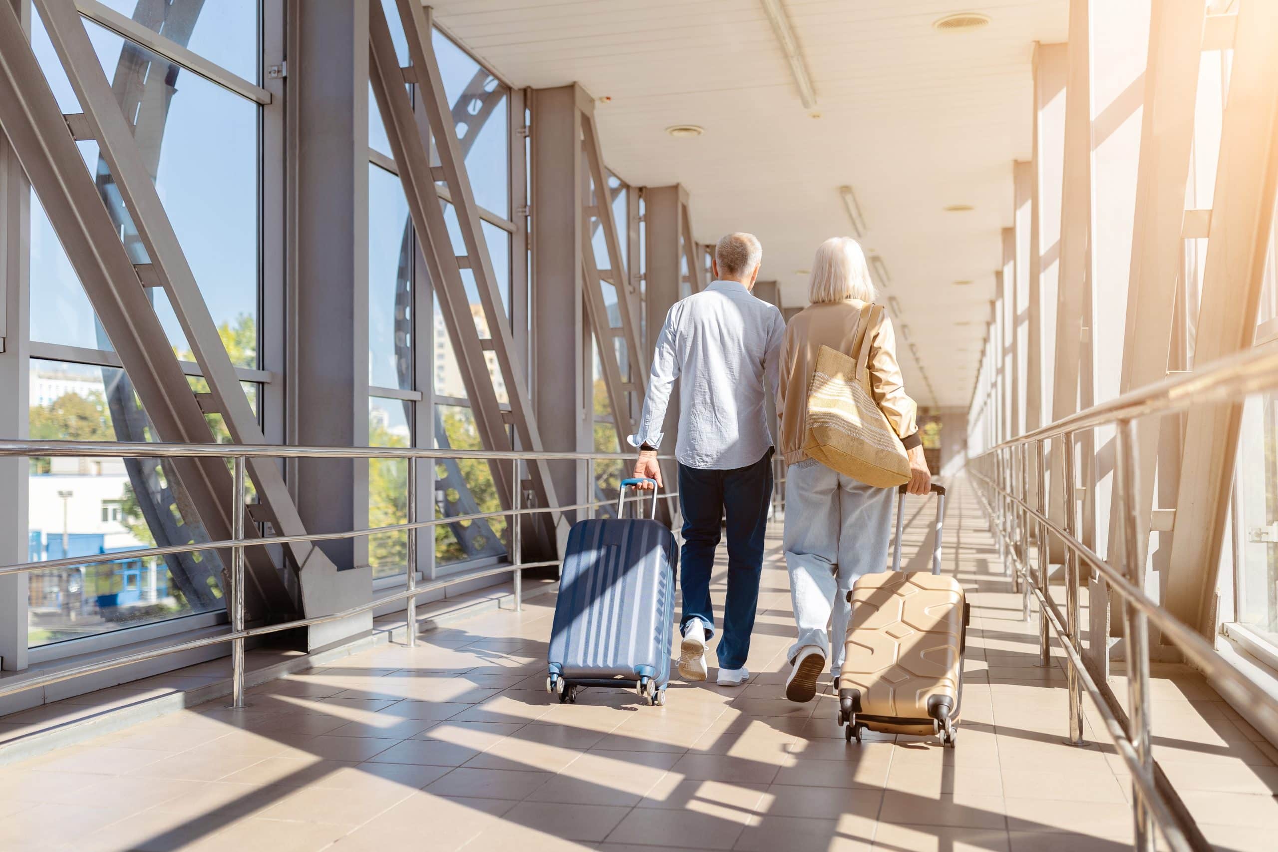 An older couple walks together down an airport corridor, each pulling a rolling suitcase. Sunlight streams through large windows as they pass by one-stop security, making their journey smoother as they head toward their destination.