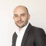 A man with a shaved head and short beard, wearing a dark suit jacket over a white shirt, stands in front of a plain light background, smiling slightly at the camera—a confident presence among world business partners.