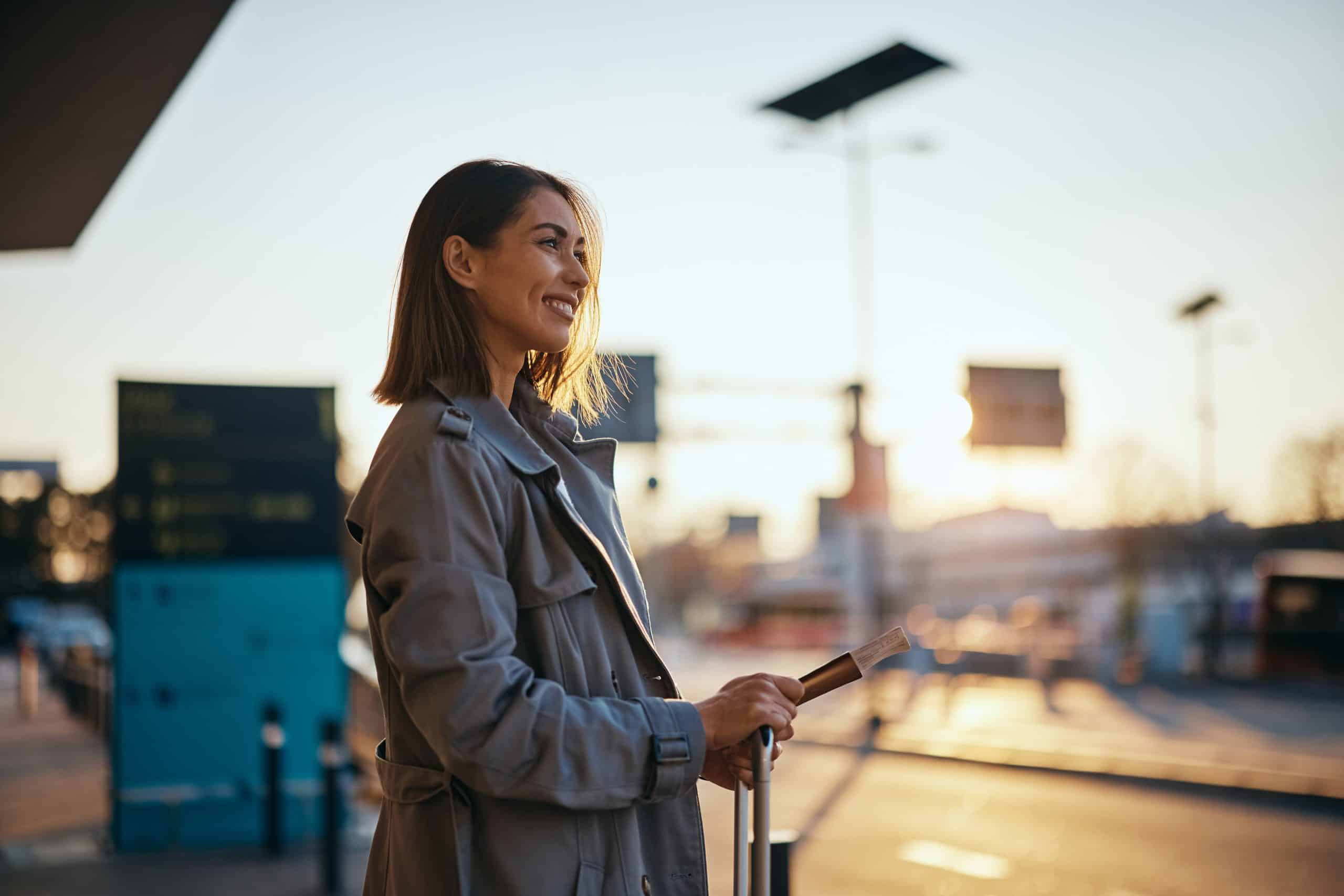 A woman in a gray coat smiles while standing outside at an airport with a suitcase and holding travel documents, celebrating her recent ASQ recognition as the sun sets in the background.