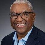 Smiling man with short gray hair and glasses, wearing a dark blazer and light blue shirt, poses against a plain gray background—reflecting the confident look of a world business partners executive.