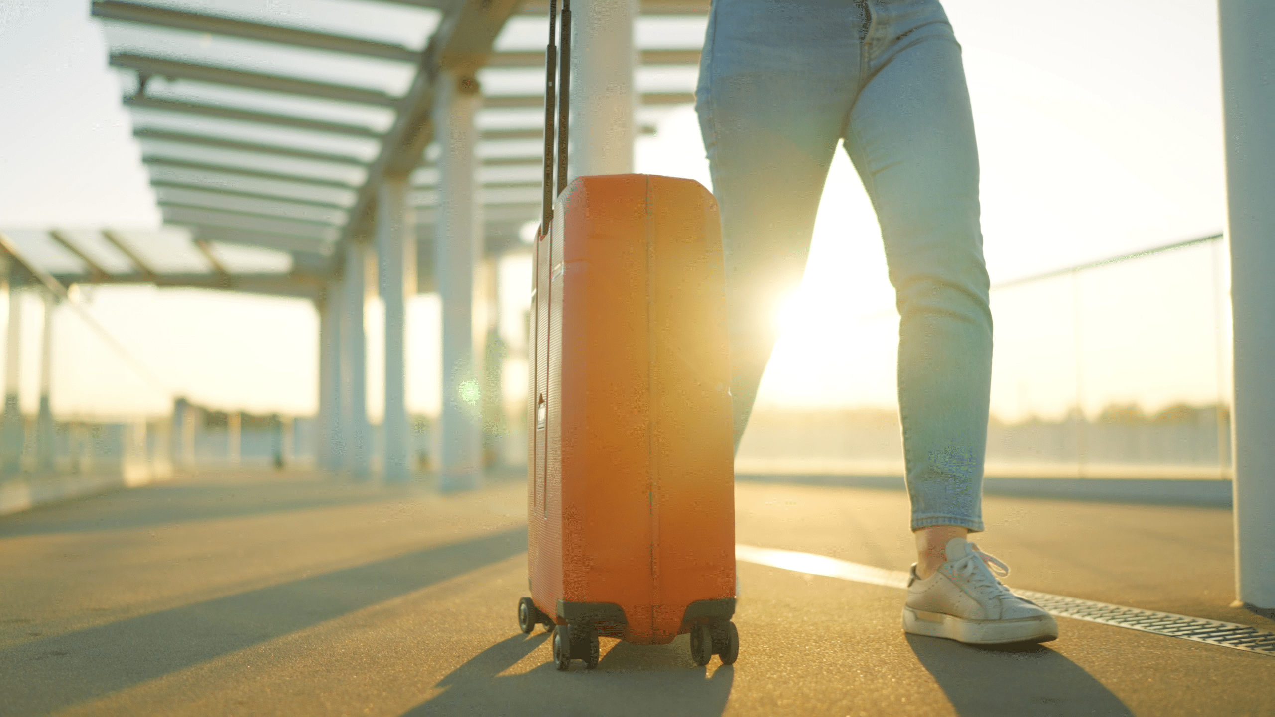 A person in jeans and sneakers walks outdoors, pulling an orange suitcase with wheels—perfect for a Front Page story. Sunlight shines in the background, suggesting early morning or late afternoon travel.