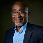 A smiling man with short hair and glasses, wearing a dark blazer, light blue checkered shirt, and blue pocket square, poses confidently in front of a dark background—ready to connect with world business partners.