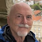 A bald man with a white mustache and goatee, wearing a blue and black jacket, smiles outdoors with historic buildings featuring red-tiled roofs blurred in the background, reflecting on connections with world business partners.