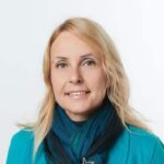 Woman with long blonde hair wearing a teal jacket and a dark blue scarf, smiling slightly, in front of a plain white background—her professional poise subtly reflecting an interest in Airport Non-Aeronautical Revenues and Activities (ANARA).