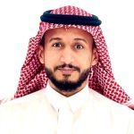 A man with a short beard wearing a red and white checkered keffiyeh and white thobe, representing the ampap alumni group, looks at the camera against a plain white background.