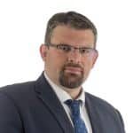 A man with short dark hair, glasses, and a beard wearing a dark suit, white shirt, and blue tie looks at the camera with a neutral expression against a white background, representing the distinguished AMPAP alumni group.