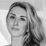 Black and white portrait of a woman with long, wavy hair, parted to the side, looking slightly upward. With defined eyebrows and a neutral expression, she appears lost in thought—an inspiring figure among the ampap alumni group. Light curtains in the background.