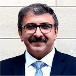 A man with short dark hair, glasses, and a mustache is smiling. He wears a dark suit, white shirt, and patterned tie, standing in front of a light brick wall—proudly representing the AMPAP alumni group.