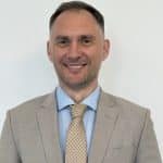 A man in a light gray suit and patterned beige tie is smiling while standing against a plain white background, embodying the professionalism often seen at Airport Non-Aeronautical Revenues and Activities (ANARA) events.