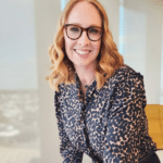 A woman with wavy blonde hair and glasses, an AMPAP alumni group member, smiles in a dark patterned blouse. She stands in a modern office space with large windows and a yellow chair in the background.
